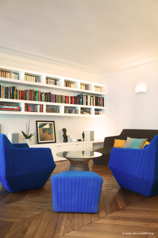 Duplex bleu et jaune. La cuisine s’ouvre sur le salon avec sa bibliothèque sur mesure et ses fauteuils contemporain du bleu qui se retrouve à plusieurs endroits de l’appartement.